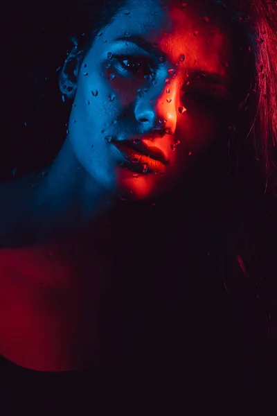 Ritratto sensuale di bella ragazza dietro vetro con gocce di pioggia con illuminazione blu rossa — Foto Stock