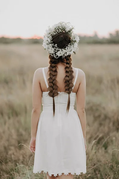 Πίσω μέρος του κοριτσιού με ένα λευκό φόρεμα σε στυλ Μπόχο με πλεξούδες και ένα λουλουδάτο στεφάνι το καλοκαίρι στο χωράφι — Φωτογραφία Αρχείου