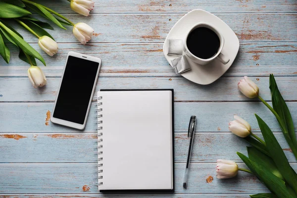 Spring blanks of a freelancer's workplace with a bouquet of white tulips, a smartphone, an empty white bouquet and a Cup of coffee in his hands on wooden background