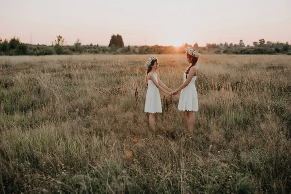 Mor och dotter tillsammans i vita klänningar med flätor och blommor kransar i boho stil i fältet sommaren vid solnedgången — Stockfoto