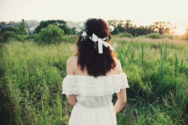 Κορίτσι με ένα λευκό φόρεμα με ένα floral στεφάνι στέκεται πίσω στο πεδίο — Φωτογραφία Αρχείου