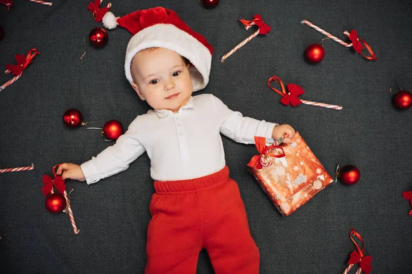 Маленький мальчик в шляпе Санты лежит среди рождественских конфет, мячей и подарочной коробки — стоковое фото