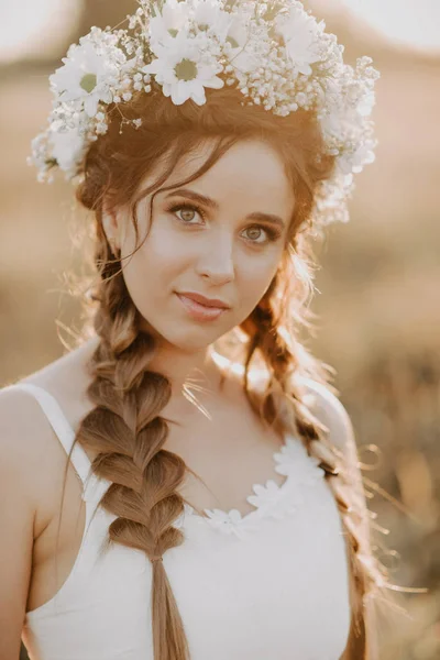 Portret dziewczyny z wieniec kwiat i warkocze w białej sukni w polu letnim o zachodzie słońca — Zdjęcie stockowe