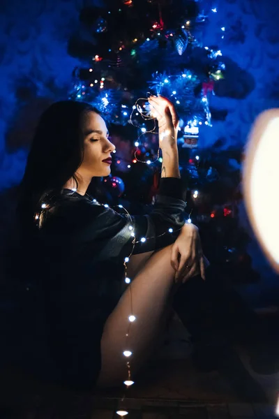 Chica bonita está sentada en el fondo de un árbol de Navidad festivo con una guirnalda —  Fotos de Stock