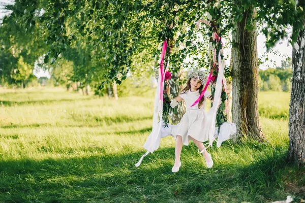 Meisje op een schommel versierd met linten en bloemen in de natuur op een zonnige zomerdag — Stockfoto