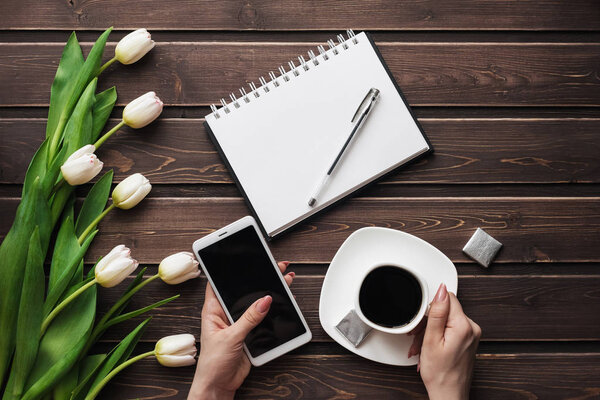 White tulips on a wooden table with an empty notebook, smartphone and a Cup of coffee in women's hands