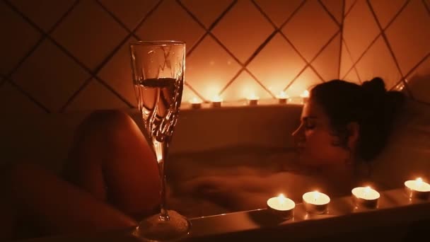 Бокал шампанского со свечами на фоне размытой расслабляющей молодой девушки в ванной комнате — стоковое видео