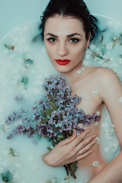 Retrato de uma bela menina desfrutando e relaxando no banho com leite e flores de maçã com um buquê de lilases — Fotografia de Stock