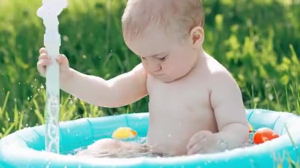 Baby Boy spelar och stänk vatten droppar i uppblåsbar pool på gräsmattan på sommaren — Stockvideo
