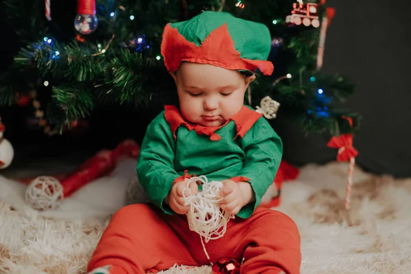 Baby boy in elf costume sitting under Christmas tree — Stock Photo, Image