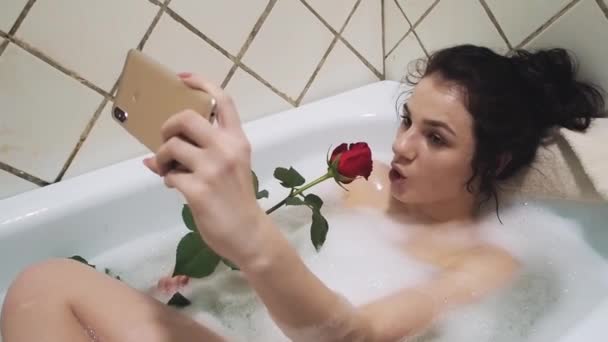 Chica divertida toma una selfie en un teléfono inteligente acostado en el baño con espuma y una rosa — Vídeos de Stock