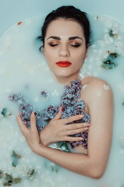 Retrato de uma bela jovem relaxante e descansando no banho com leite e flores de lilás — Fotografia de Stock
