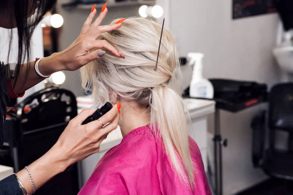 Mãos de um cabeleireiro profissional fazer um penteado baixo bun para uma menina loira — Fotografia de Stock