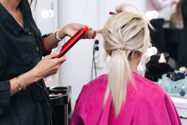 Mãos de um cabeleireiro com um ferro fazer o cabelo de uma menina loira — Fotografia de Stock