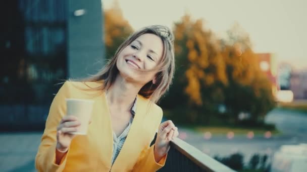 Femme vêtue d'une veste jaune buvant du café et souriant à la ville . — Video