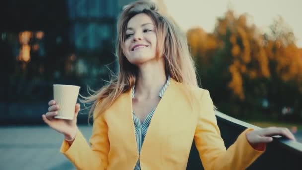 Mulher vestida de casaco amarelo bebendo café e sorrindo para a cidade . — Vídeo de Stock