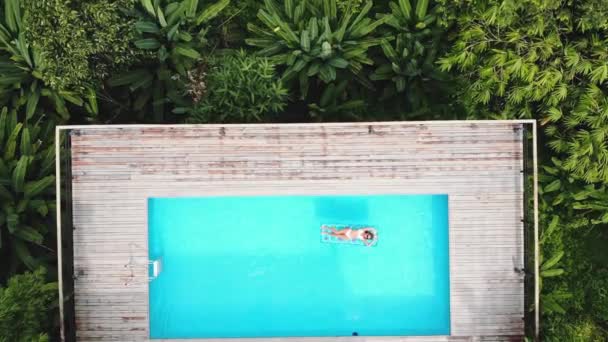 AERIAL. Top view. Camera zooming. Outdoor lifestyle portrait of young pretty sexy girl posing with neon bright swimming mattress at the pool in luxury villa at the jungle, wearing bikini and — Stock Video