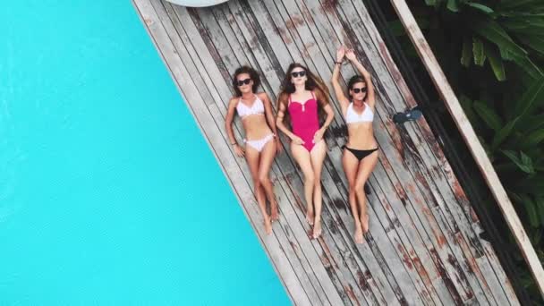 AERIAL. Cámara alejándose. Vista superior de tres jóvenes con gafas de sol, tumbadas cerca de la piscina en la lujosa villa de la selva. Tres amigos relajándose en la piscina . — Vídeos de Stock