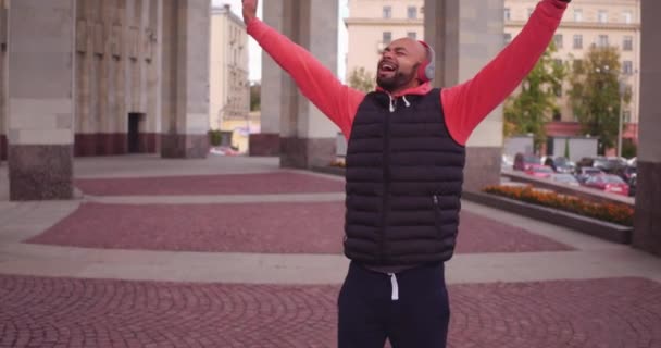 Portrait extérieur de positif afro-américain buvant du café, dansant et souriant à la caméra tout en marchant dans la rue, portant une tenue à la mode et des écouteurs . — Video