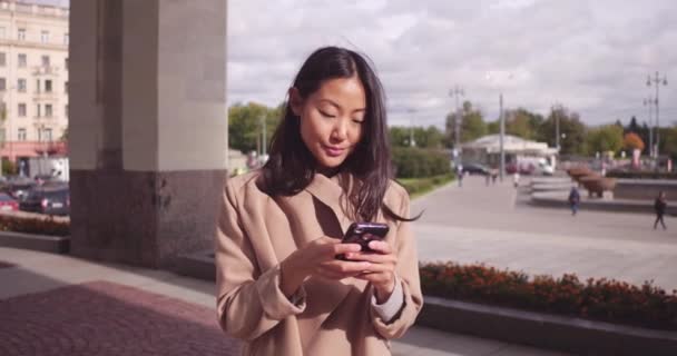 Phone Asian business woman texting sms on cellphone app in city street, urban lifestyle. Europe travel vacation tourist using smartphone outside. Chinese businesswoman in red trench coat. — Stock Video