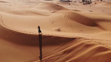 AERİAL. Dubai çölünde gün batımında kumulda yürüyen uzun elbiseli bir kadın.