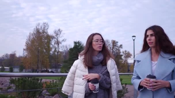 Outdoors fashion portrait of two cheerful girls, asian and arabian drinking coffee. Walking on the embankment. — Stock Video