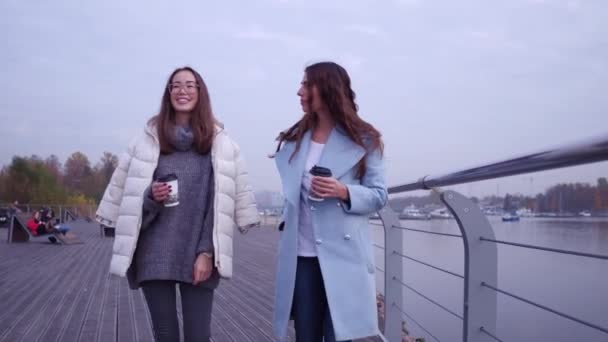 Outdoors fashion portrait of two cheerful girls, asian and arabian drinking coffee. Walking on the embankment. — Stock Video