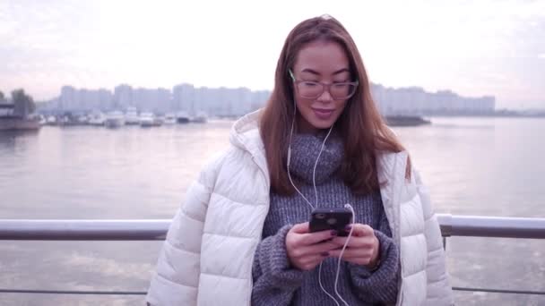 Mujer asiática joven usando teléfono inteligente de pie en la costa del mar de terraplén. Mujer vestida con suéter gris y chaqueta blanca sosteniendo dispositivo móvil sobre fondo de agua abierta, primer plano . — Vídeo de stock