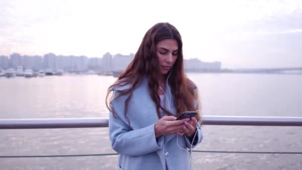 Close up retrato filmado de mulher bonita em casaco azul em pé no dique, olhando para a exibição de seu smartphone sorrindo e rindo alegremente, impressionado com o conteúdo de mídia da web . — Vídeo de Stock