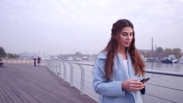 Alegre casualmente vestida mujer en azul abrigo mensajes de texto en el teléfono inteligente mientras camina en el terraplén de la ciudad, sonriendo . — Vídeos de Stock