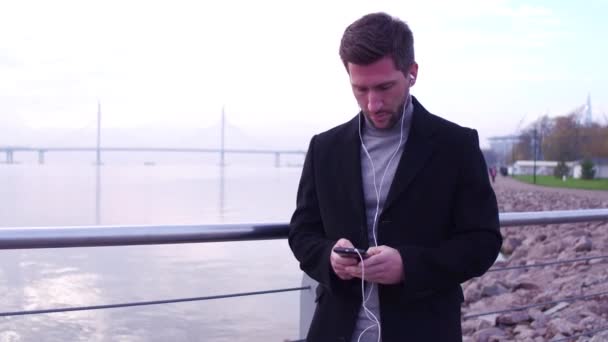 イヤホンの美しい背景を望む都市堤防の上に立って、電話チャットでハンサムな男. — ストック動画
