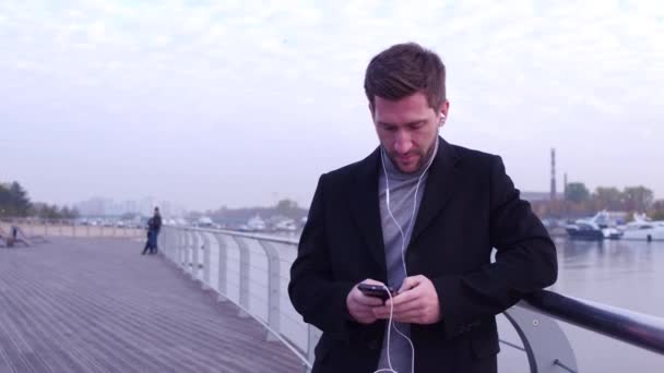 En kjekk mann med øretelefoner som chatter over telefon, som står på byens voll med vakker bakgrunn. . – stockvideo