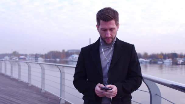 Seriöser Mann mit Kopfhörern, der telefoniert, auf dem Stadtdamm mit schönem Hintergrundblick läuft. — Stockvideo