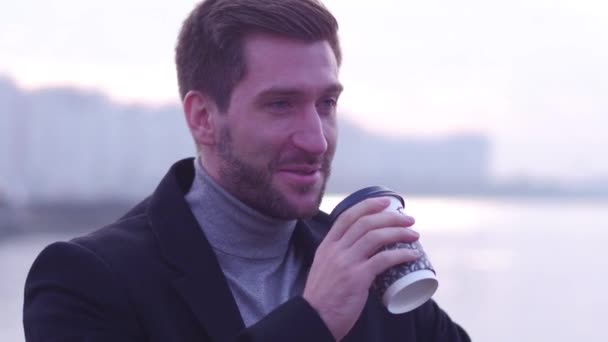 Jeune homme souriant, dehors - dehors. Gros plan du mâle en manteau noir, froidement le matin, buvant du café sur le remblai . — Video