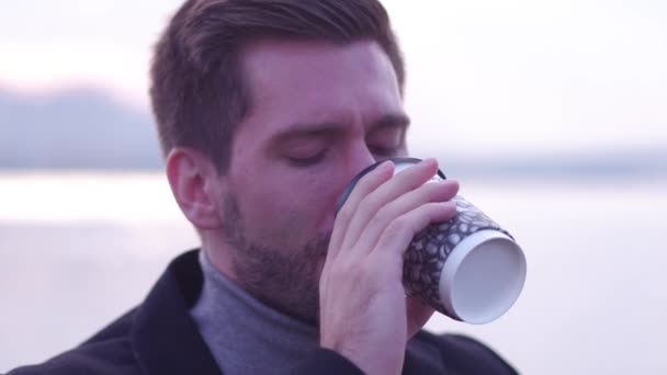 严肃的年轻人, 户外。穿着黑色外套的男性的特写, 寒冷的早晨, 在堤防上喝咖啡. — 图库视频影像