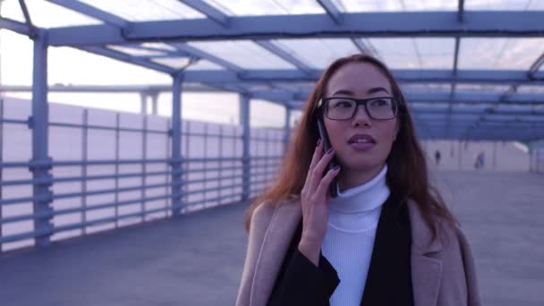 Teknik. Stadsvandring. Kvinna porträtt. Vacker ung asiatisk kvinna i glasögon talar på mobiltelefonen och ler medan promenader utomhus. — Stockvideo