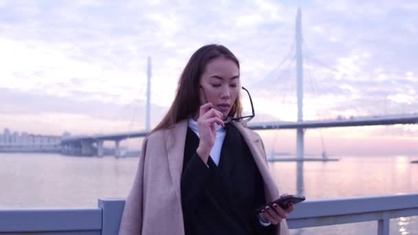 Fiatal gyönyörű ázsiai businesswomanin szemüveg segítségével a smartphone, a gyönyörű naplemente háttér első városi híd. — Stock videók