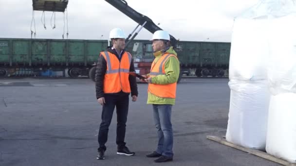 Dua insinyur yang memakai topi keras berbicara bersama saat berdiri di atas tempat latihan komersial yang besar melacak persediaan — Stok Video