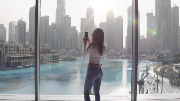 Mujer joven tomando fotos en la ventana grande y mirando a los edificios de la ciudad de Dubai. Puesta de sol. — Vídeos de Stock