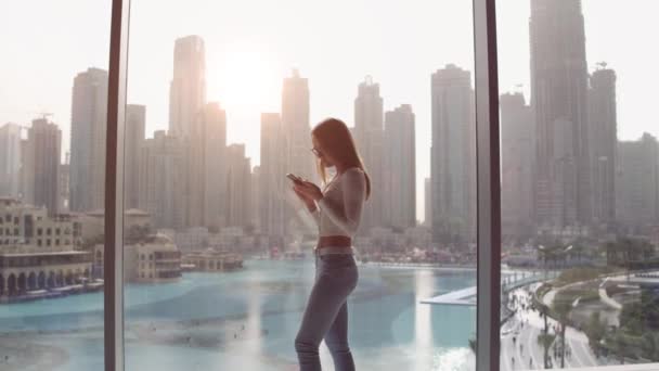 Jovem mulher digitando ao telefone na grande janela e olhando para os edifícios da cidade de Dubai. Pôr do sol . — Vídeo de Stock