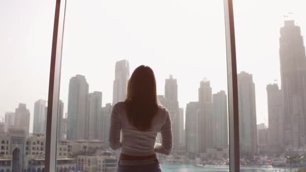 Mulher jovem tirando fotos na grande janela e olhando para os edifícios da cidade de Dubai. Pôr do sol. — Vídeo de Stock
