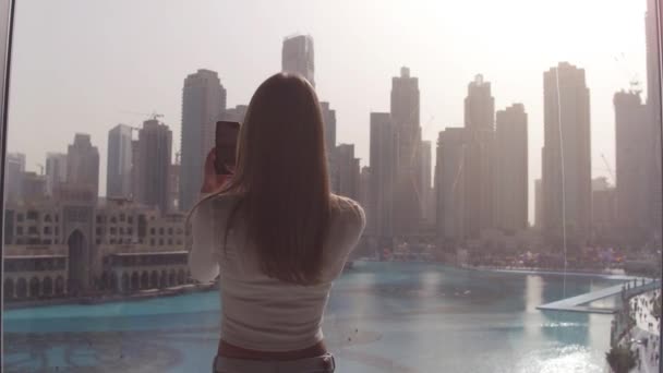 Jovem mulher digitando ao telefone na grande janela e olhando para os edifícios da cidade de Dubai. Pôr do sol . — Vídeo de Stock
