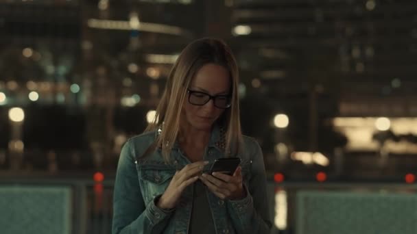 女性はビジネスタワーの上の夜の時間に街のスマートフォンで働く。青墨色の背景. — ストック動画