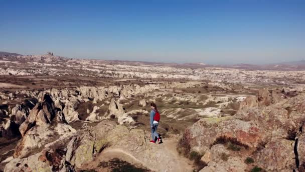 AERIAL. Cámara moviéndose alrededor de mujer joven levantando las manos en la montaña por la mañana. Viajero con mochila roja de pie en la montaña por la mañana . — Vídeos de Stock