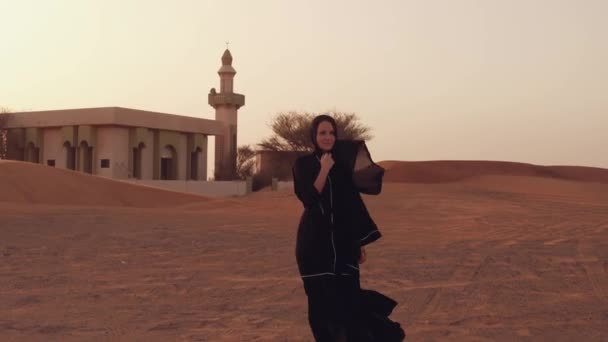 Portrait of a young Arab woman wearing traditional black clothing during beautiful sunset over the desert. Mosque on the background — Stock Video