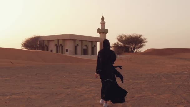 Porträt einer jungen Araberin in traditioneller schwarzer Kleidung bei schönem Sonnenuntergang über der Wüste. Moschee im Hintergrund — Stockvideo