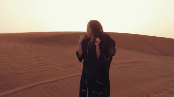 Portret van een jonge Arabische vrouw in traditionele zwarte kleding tijdens een prachtige zonsondergang boven de woestijn. — Stockvideo