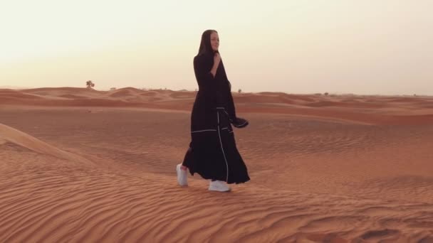 Portret van een jonge Arabische vrouw in traditionele zwarte kleding tijdens een prachtige zonsondergang boven de woestijn. — Stockvideo