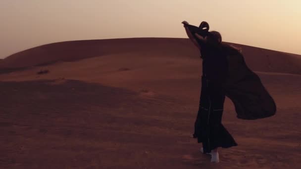 Retrato de una joven árabe vestida con ropa negra tradicional durante el hermoso atardecer sobre el desierto. — Vídeos de Stock