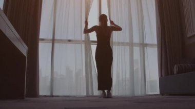 Perdeleri açan ve sabahları pencereden şehir manzarasına bakan kadının arka görüntüsü. Dubai şehrindeki modern binalar